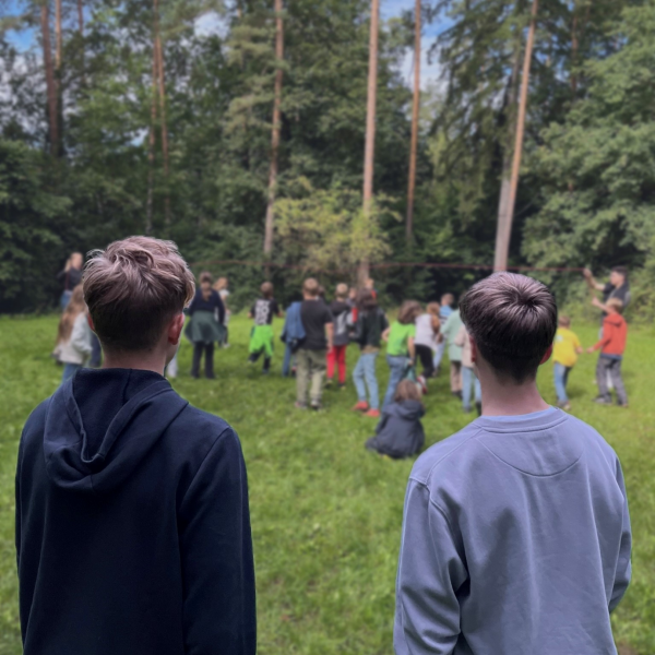 Zwei Jugendliche beobachten eine Gruppe von Kindern, die auf einer Wiese im Wald gemeinsam ein Spiel spielen