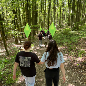 Jugendliche laufen einen Waldweg entlang; über einigen Köpfen sind stilisierte Spielsymbole wie aus einem Videospiel eingeblendet