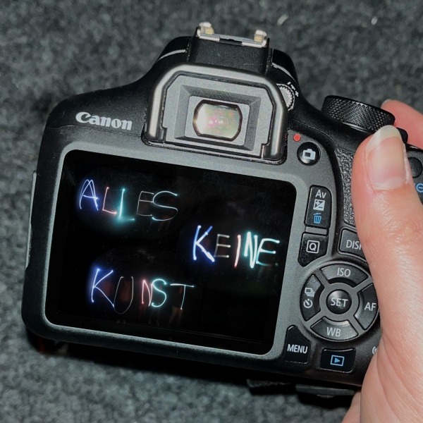 Digitalkamera von hinten, auf dem Display steht in bunten Lichtbuchstaben "Alles keine Kunst"
